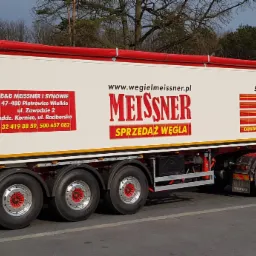 Ciężarówka z naczepą firmy Meissner z reklamą sprzedaży węgla, drewna, kruszyw i piasku, zaparkowana na parkingu z widocznym budynkiem w tle.