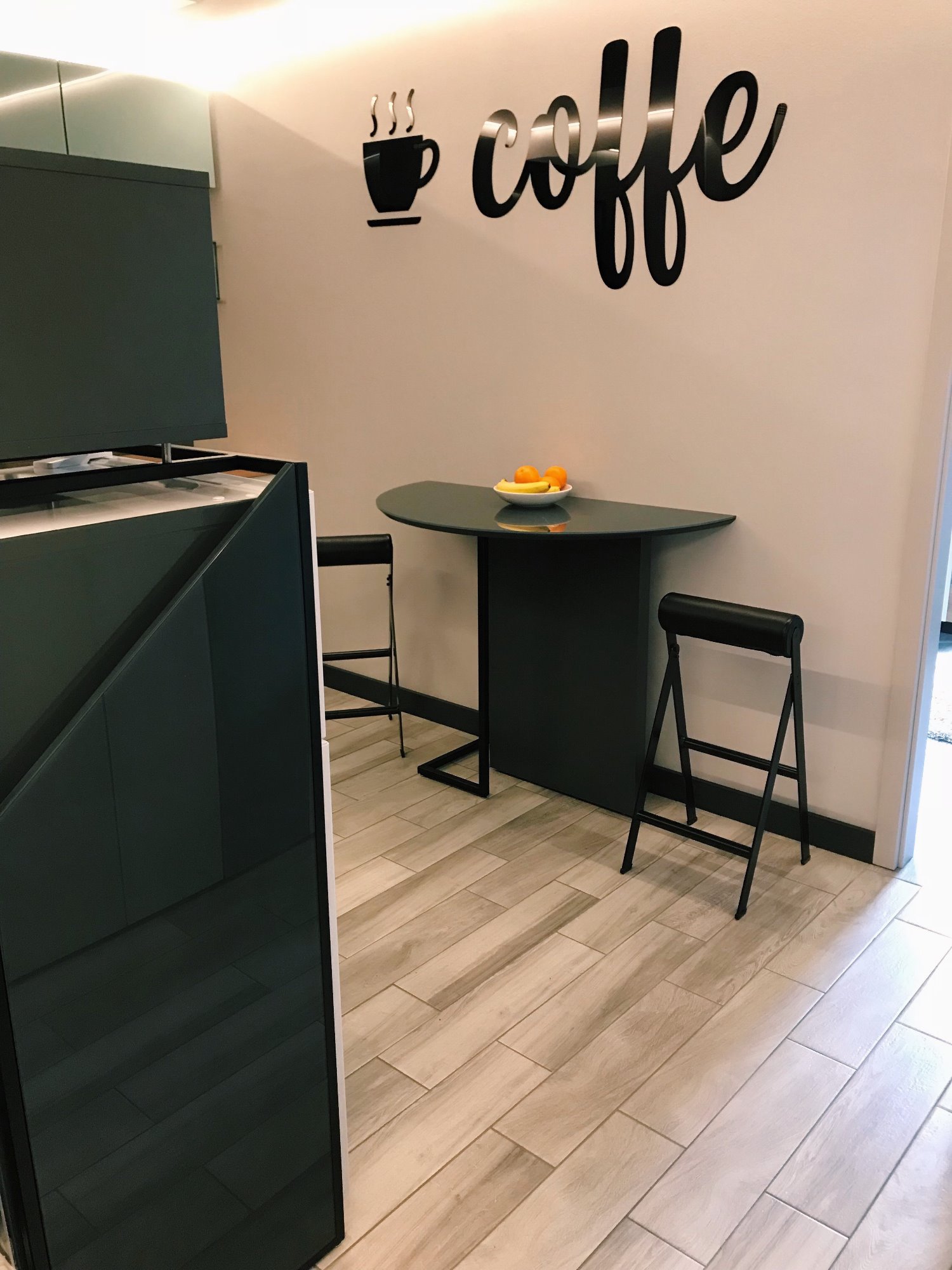 Częściowy widok nowoczesnej kuchni z blatem i dwoma składanymi stołkami barowymi, na blacie misa z pomarańczami, na ścianie dekoracja 'coffe' i motyw filiżanki, podświetlenie LED.