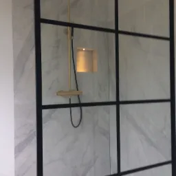 Kabina prysznicowa typu walk-in z czarnymi profilami i złotą armaturą na tle marmurowych płytek.