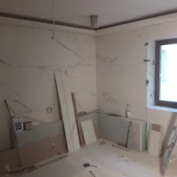 Łazienka w trakcie prac: ściany wyłożone białymi płytami imitującymi marmur, widoczne narzędzia, drabina, butelka z płynem i otwór w ścianie pod instalację.