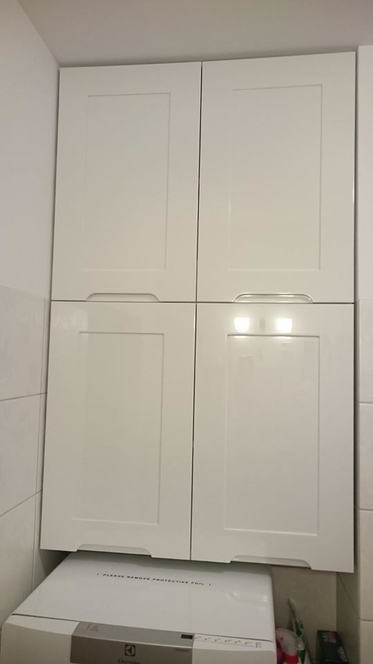 Białe, lakierowane szafki kuchenne wiszące nad pralką Electrolux w łazience z białymi płytkami.