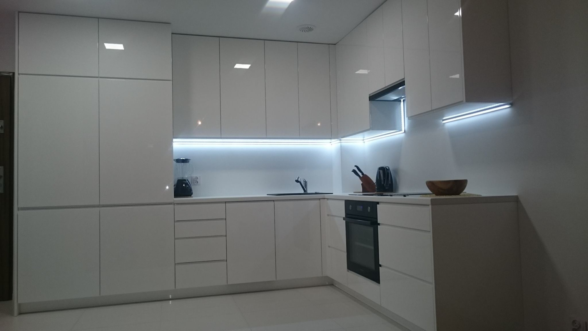 Nowoczesna, minimalistyczna kuchnia z białymi, lakierowanymi frontami, oświetleniem LED pod szafkami wiszącymi i sprzętami AGD w zabudowie.