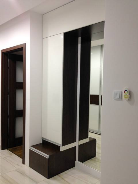 Nowoczesna szafa wnękowa na wymiar z białymi frontami i ciemnobrązowymi akcentami, z siedziskiem w dolnej części, z widocznym wyłącznikiem światła na ścianie po prawej stronie.