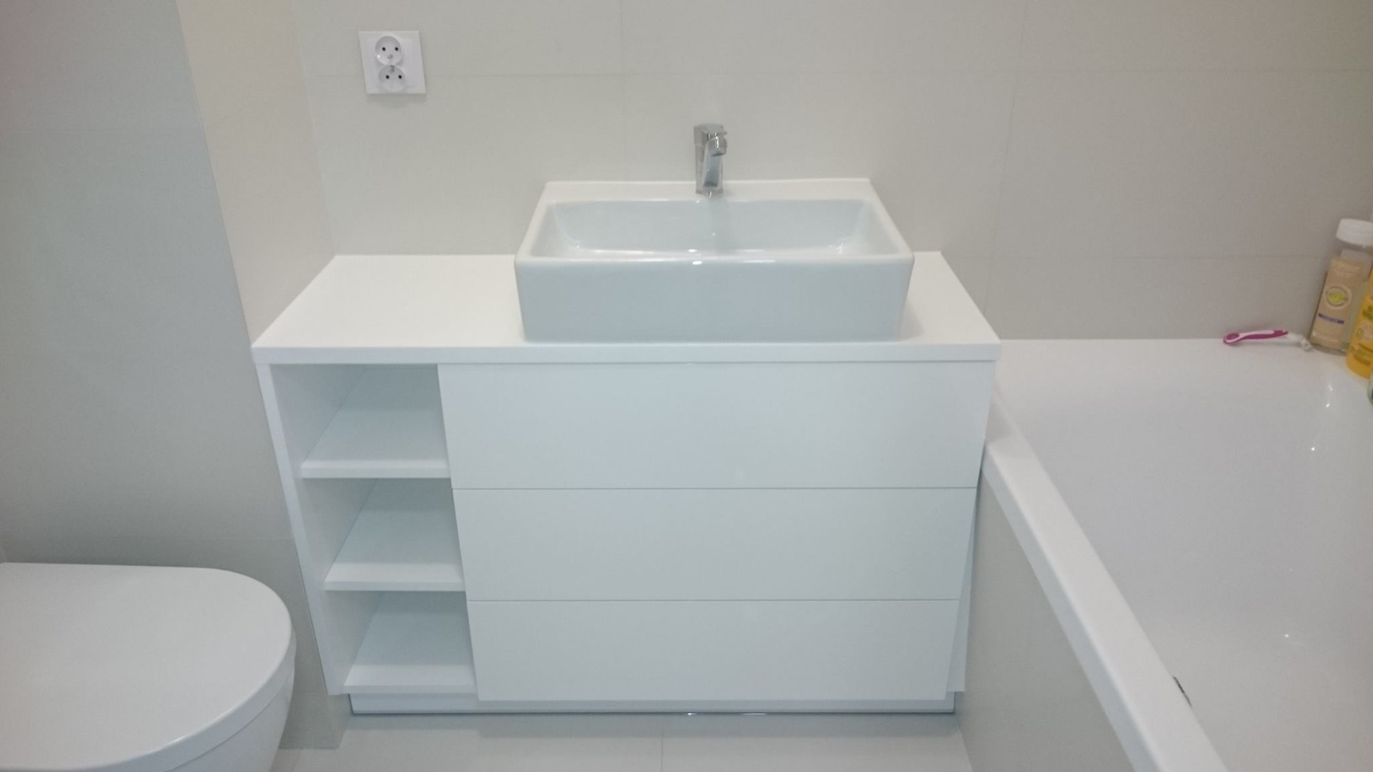 Biała szafka łazienkowa z trzema szufladami i otwartymi półkami, na niej kwadratowa umywalka nablatowa z chromowaną baterią, obok fragment wanny i toalety w minimalistycznej łazience.