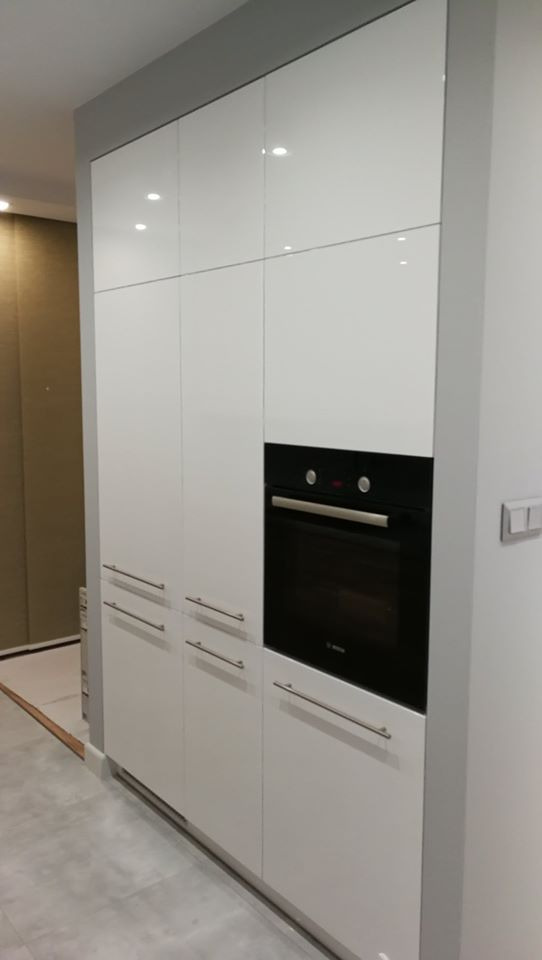 Wysoka, zabudowana szafa kuchenna z białymi, lakierowanymi frontami i wbudowanym piekarnikiem Bosch, widoczna na tle szarej ściany i podłogi z płytek.