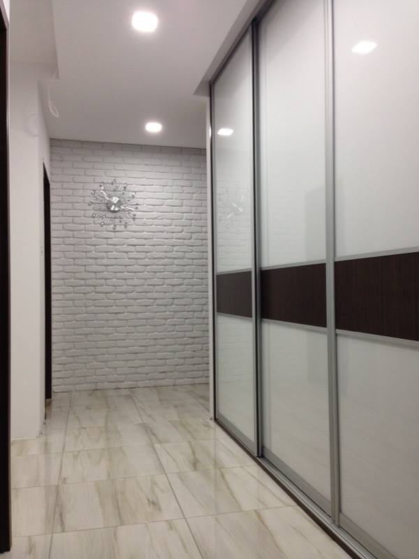 Szafa przesuwna z białymi frontami i brązowym pasem dekoracyjnym, zamontowana w korytarzu z dekoracyjną ścianą z białej cegły i zegarem w Łyse.