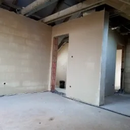 Świeżo otynkowane ściany poddasza z widoczną konstrukcją drewnianą dachu, otwory na gniazdka elektryczne i przewody na podłodze. W tle widoczne narzędzia budowlane.