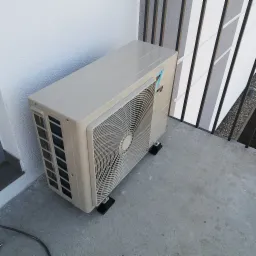 Zewnętrzna jednostka klimatyzacji zamontowana na balkonie, widok z góry, widoczne czarne podkładki pod urządzeniem i fragment kabla zasilającego.