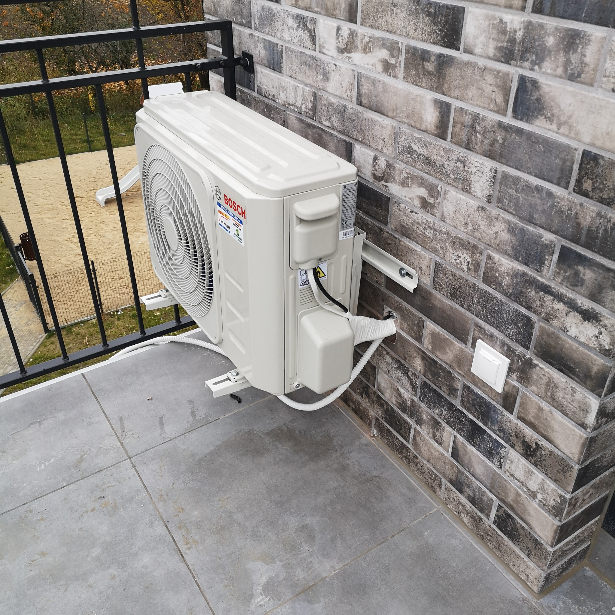 Biały klimatyzator marki Bosch zamontowany na zewnątrz na balkonie, widoczna elewacja z cegły klinkierowej i barierka balkonu.