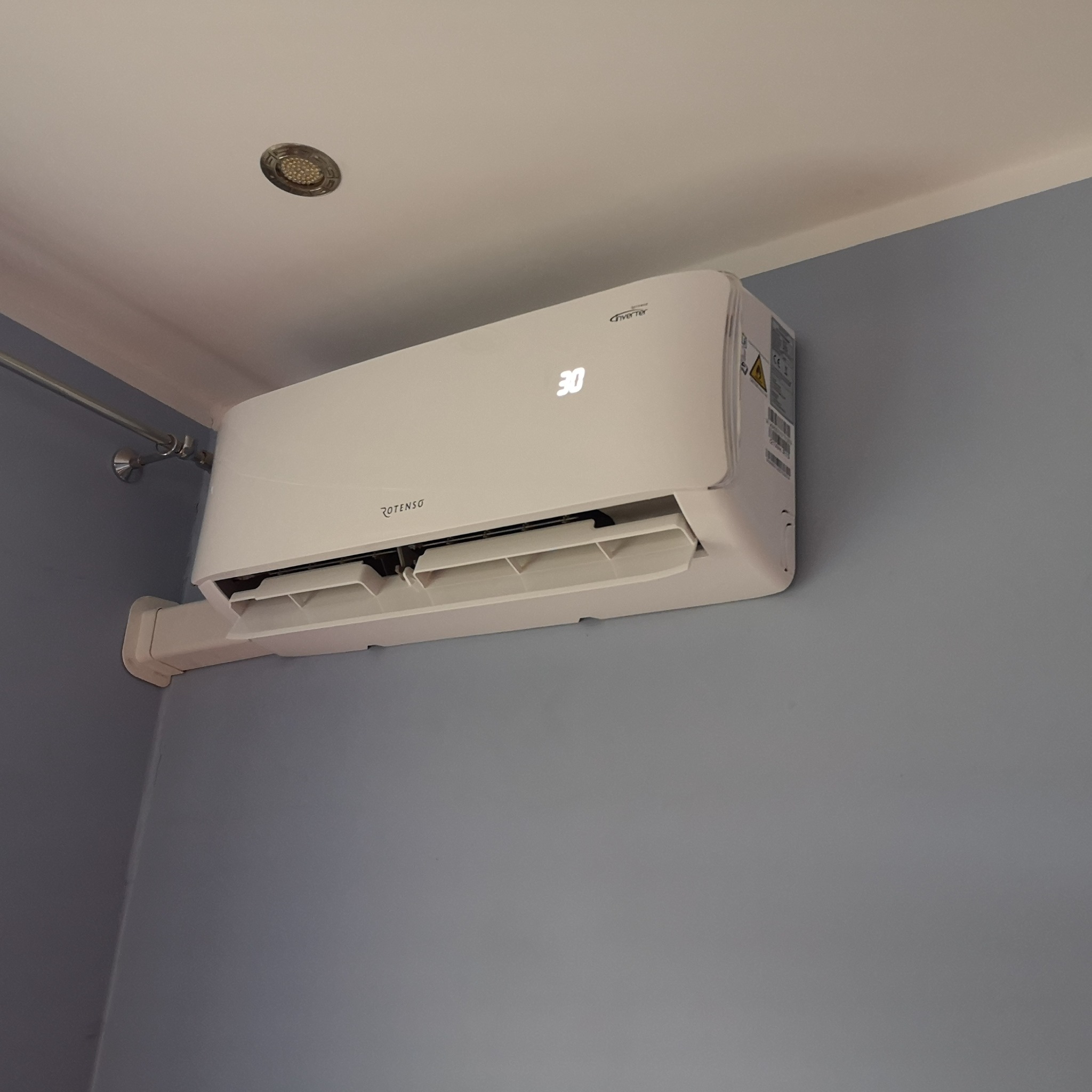 Biały klimatyzator Rotenso z wyświetlaną temperaturą 30 stopni Celsjusza, zamontowany na szarej ścianie w pomieszczeniu z widocznym oświetleniem sufitowym.