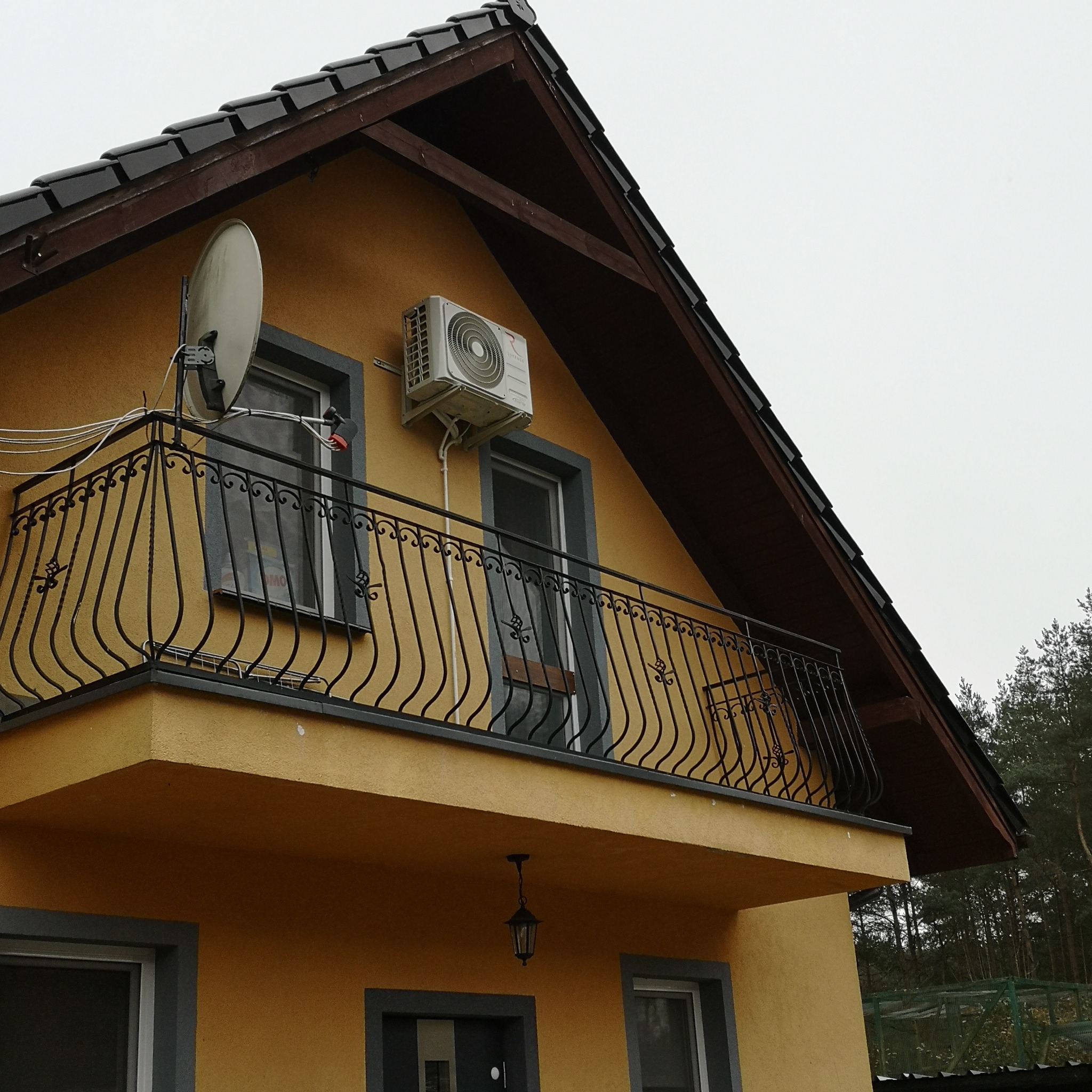 Żółta elewacja domu z balkonem i zamontowaną jednostką zewnętrzną klimatyzacji oraz anteną satelitarną.