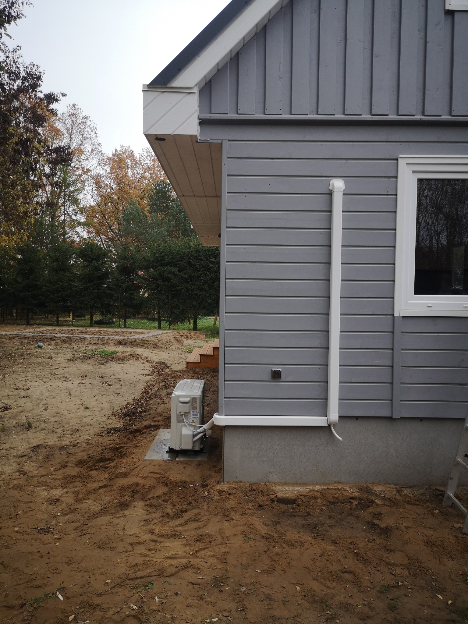 Zewnętrzna jednostka klimatyzacji zamontowana przy szarym, drewnianym domu z widocznymi rurami odprowadzającymi skropliny, na tle piaszczystego terenu.