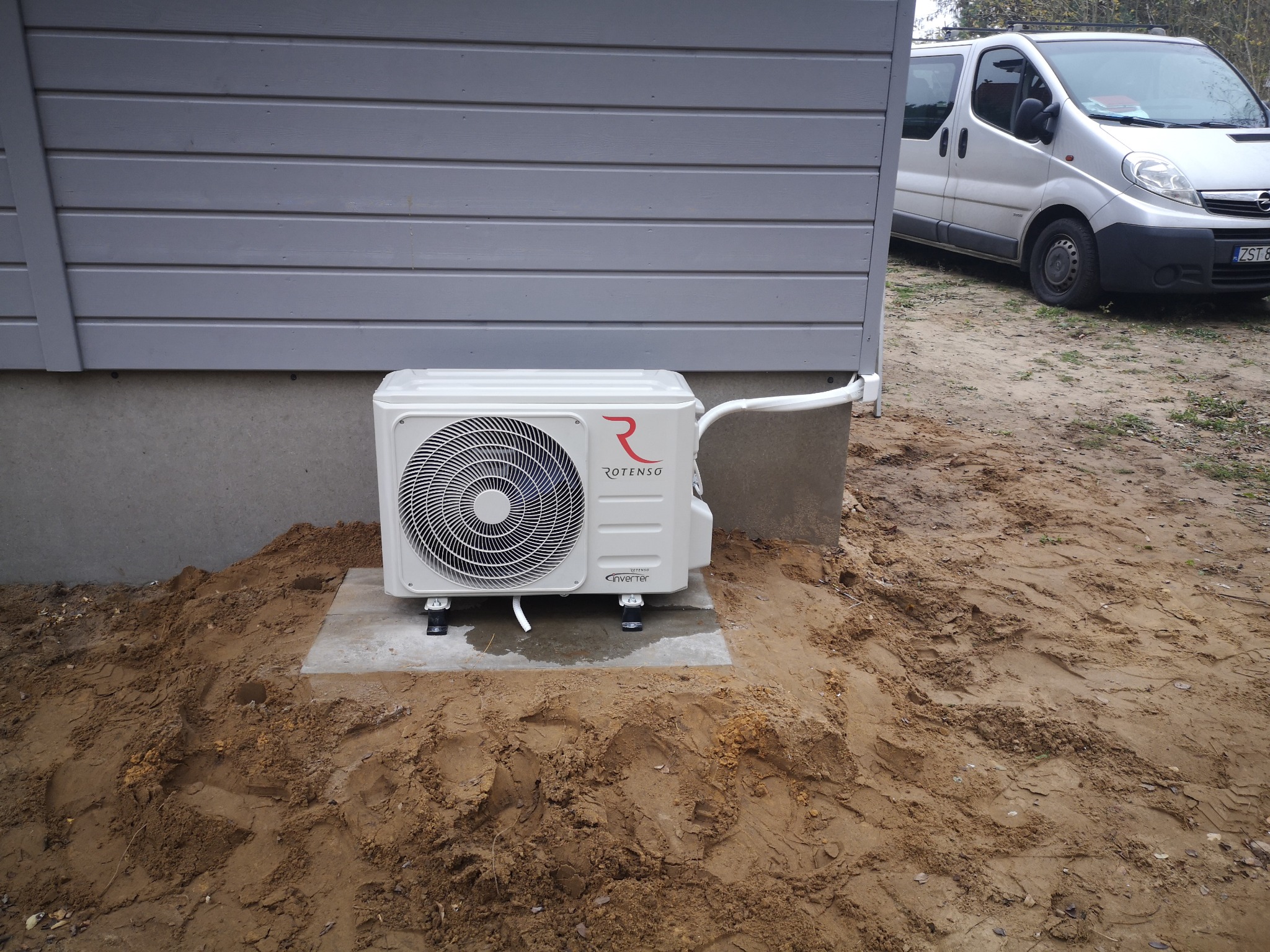 Zewnętrzna jednostka klimatyzacji Rotenso zamontowana przy szarej ścianie budynku na betonowej podstawie, otoczona ziemią; w tle zaparkowany srebrny van.