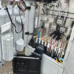 Otwarta szafka telekomunikacyjna z licznymi kablami światłowodowymi oznaczonymi kolorami, splatarką światłowodową Fitel S179 oraz otwartą walizką narzędziową u dołu.