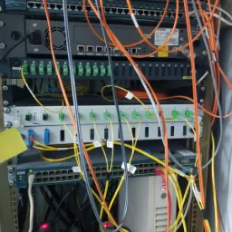 Szafa rack z zainstalowanymi urządzeniami sieciowymi, widoczne patch panele, przełączniki Cisco Catalyst, oraz liczne kable światłowodowe i ethernetowe w kolorach pomarańczowym, żółtym i szarym,...