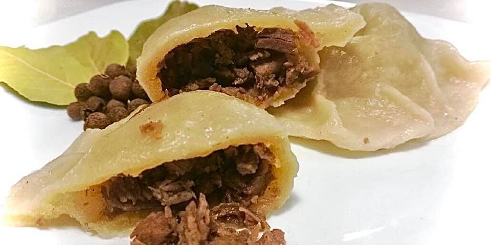 Dwa przekrojone pierogi z mięsnym farszem na białym talerzu, obok liść laurowy i ziele angielskie.