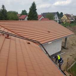 MWer & Co Sp. z o.o. - Montaż rynny na nowym dachu z dachówki ceramicznej w kolorze ceglastym, widoczny pracownik w odblaskowej kurtce i fragment budynku z bloczków.