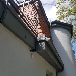 Szara obróbka blacharska dachu z balkonem, widoczny system rynnowy i dachówka ceramiczna w kolorze ceglastym, na tle budynku o białej elewacji i zieleni drzew.