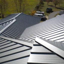 Widok z góry na nowo położony, metalowy dach z blachy trapezowej w kolorze srebrnym, z widocznymi elementami łączeń i załamań, na tle zielonego terenu z drzewami i zaparkowanym białym busem.