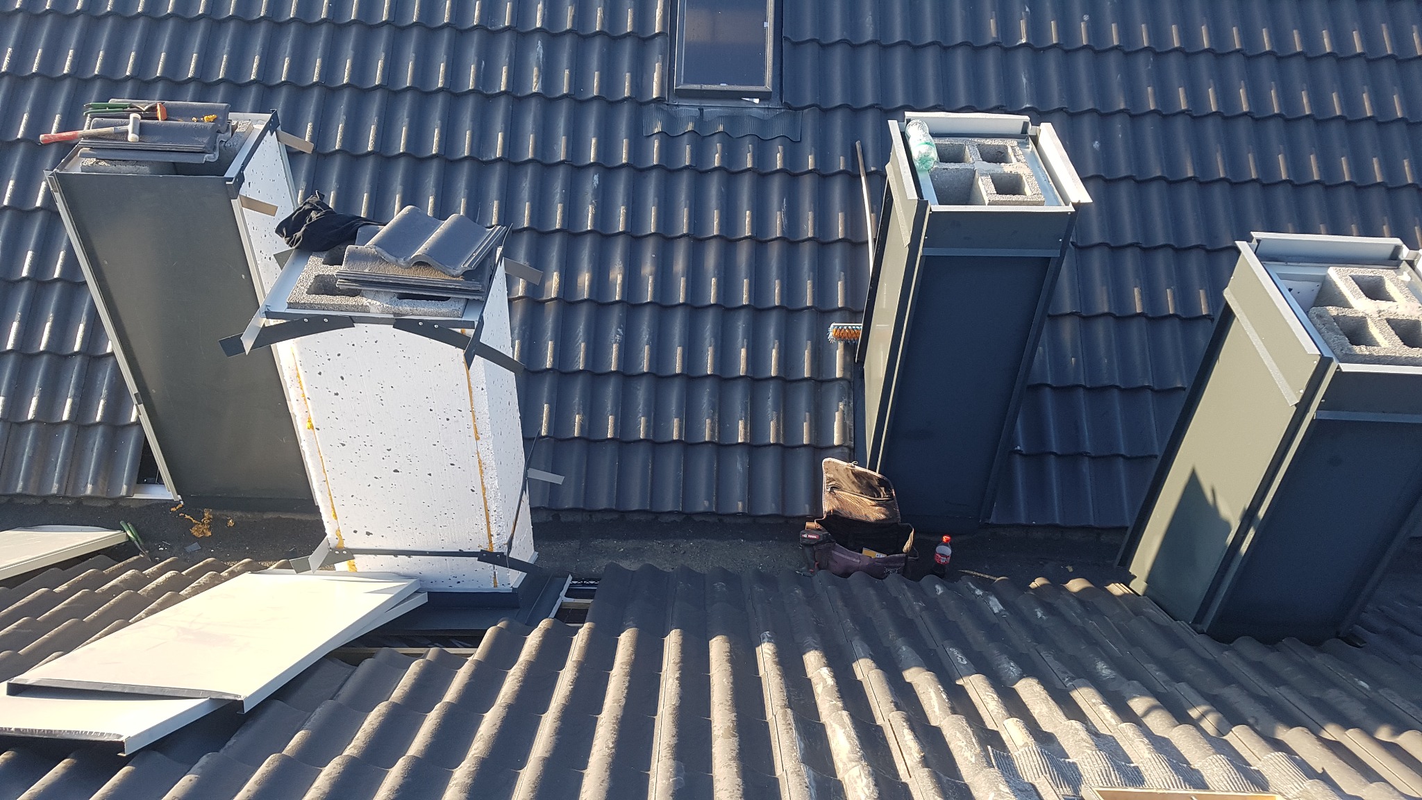 Obróbka blacharska kominów z blachy na dachu pokrytym szarą dachówką karpiówką, widoczne narzędzia i materiały dekarskie.