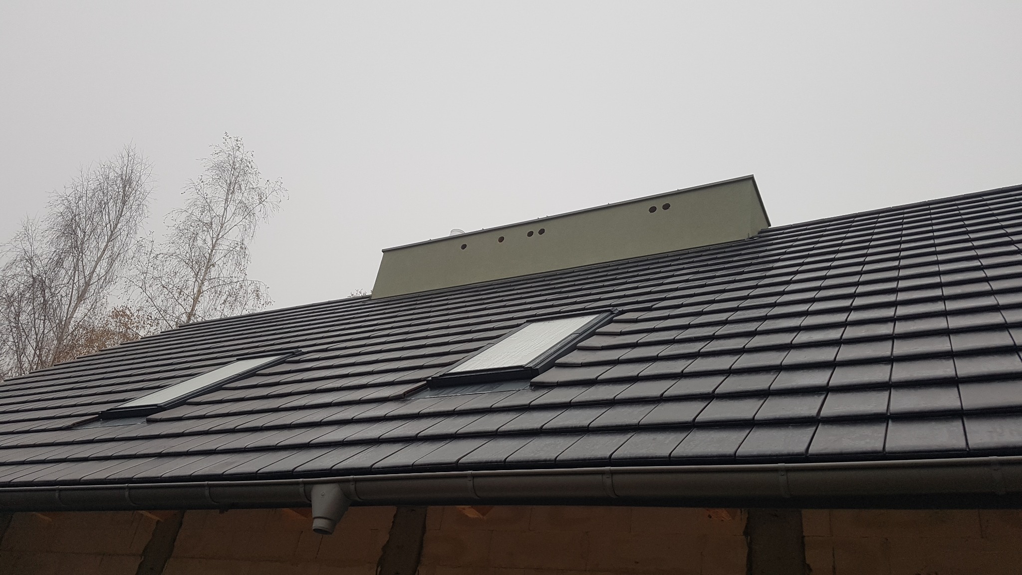 Nowy dach pokryty ciemną dachówką ceramiczną z dwoma oknami dachowymi, widok na komin wentylacyjny i nagie drzewa w tle, pochmurne niebo.