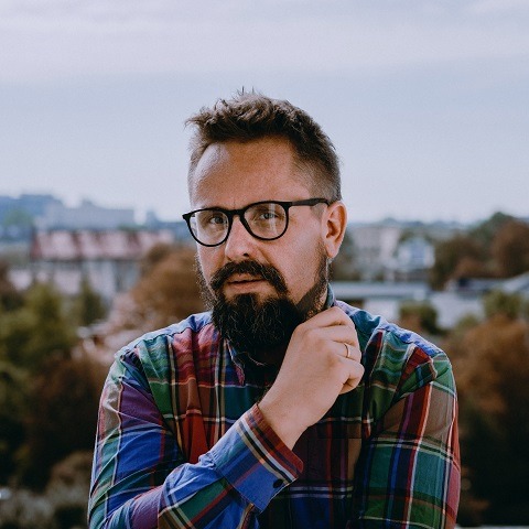 Portret mężczyzny w okularach i kolorowej koszuli w kratę, z brodą, stojącego na balkonie z widokiem na miasto w tle.