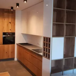 Nowoczesna kuchnia z drewnianymi frontami szafek o wyraźnym usłojeniu, białymi szafkami wiszącymi i otwartym regałem z elementami białymi, oświetlona punktowymi lampami sufitowymi.