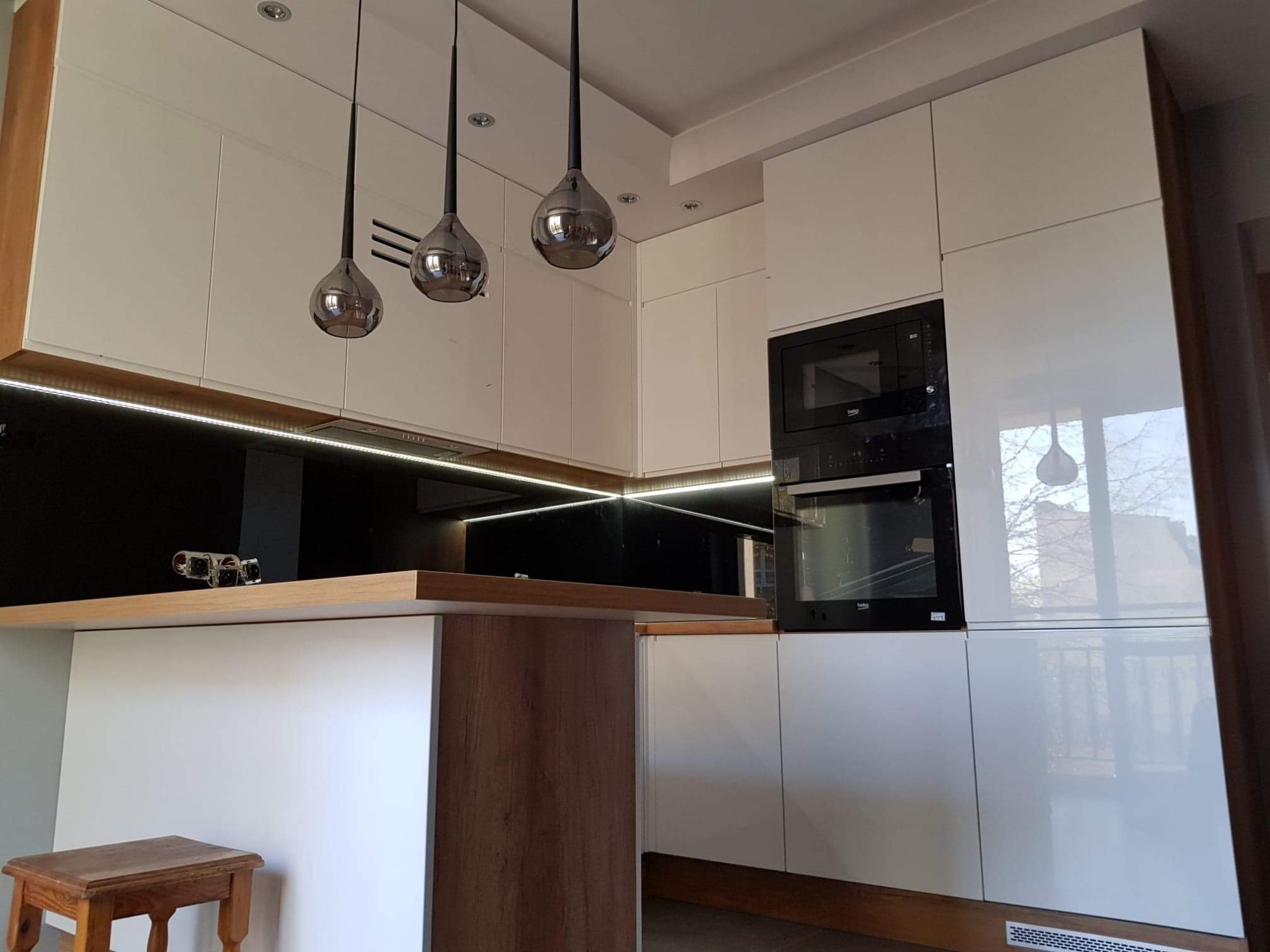 Nowoczesna kuchnia z białymi, lakierowanymi frontami, drewnianym blatem i trzema wiszącymi lampami o kulistych kloszach, oświetlenie LED pod szafkami.