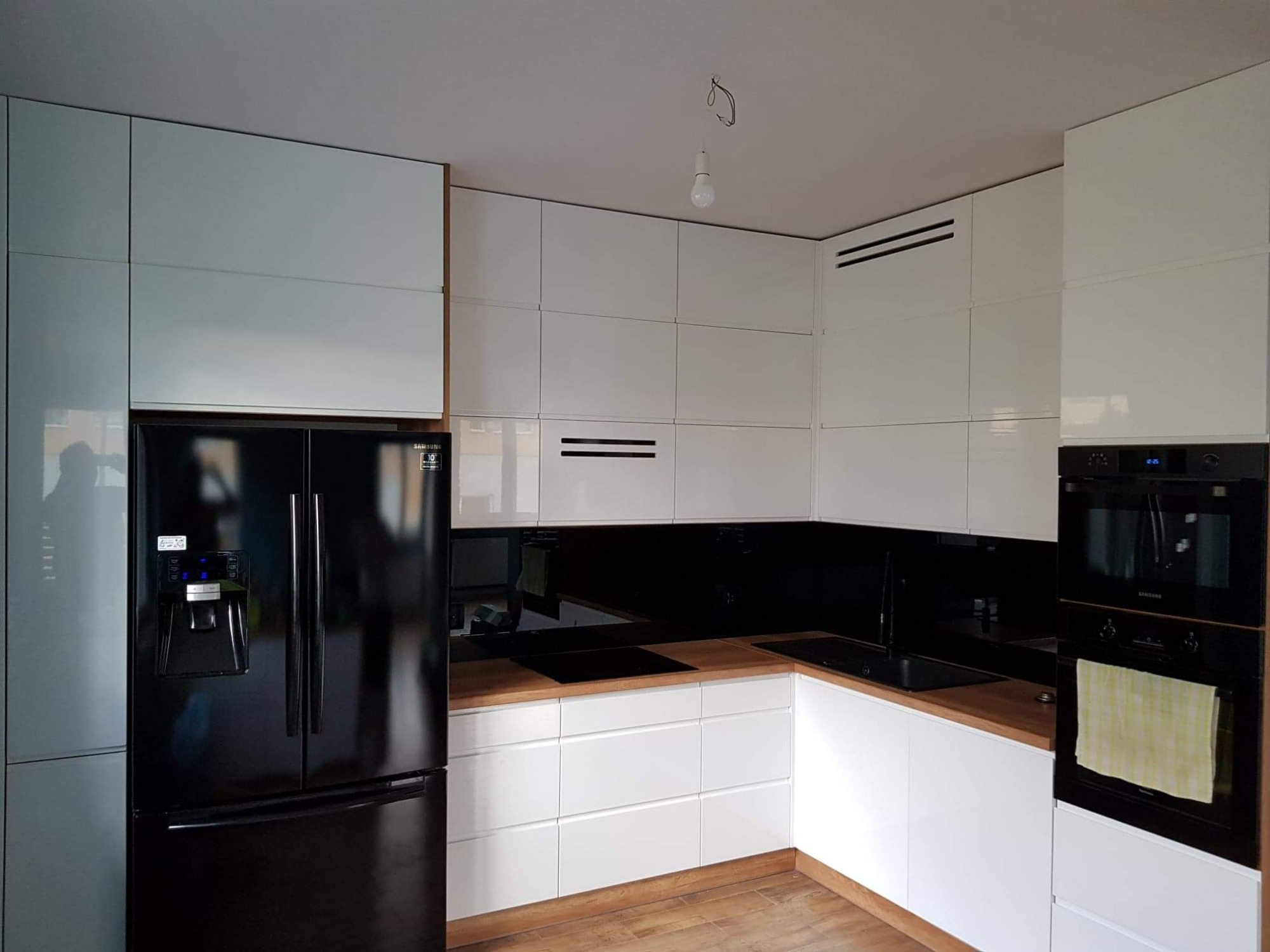 Nowoczesna kuchnia z białymi, lakierowanymi frontami szafek, drewnianym blatem i czarnym panelem szklanym nad blatem, z czarną lodówką Samsung i piekarnikiem w zabudowie.