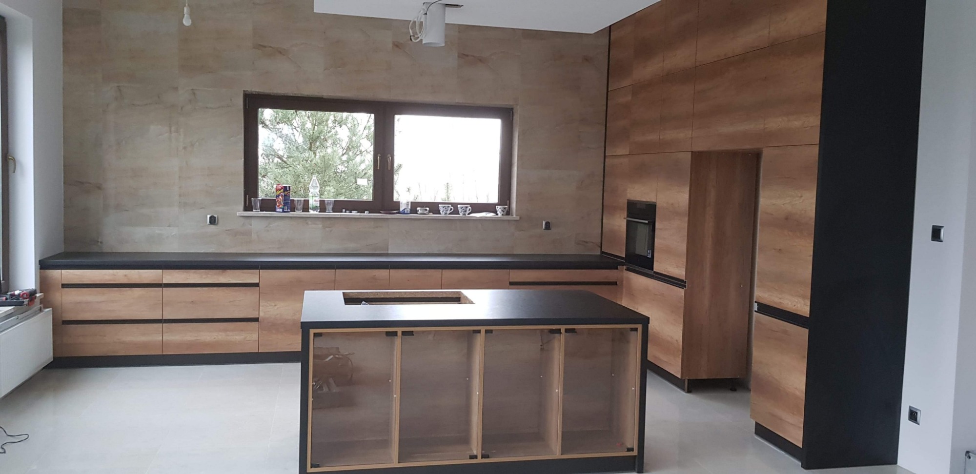 Wnętrze kuchni w trakcie montażu, z drewnianymi szafkami i czarnymi blatami, wyspa kuchenna w surowej ramie, okno z widokiem na drzewa.