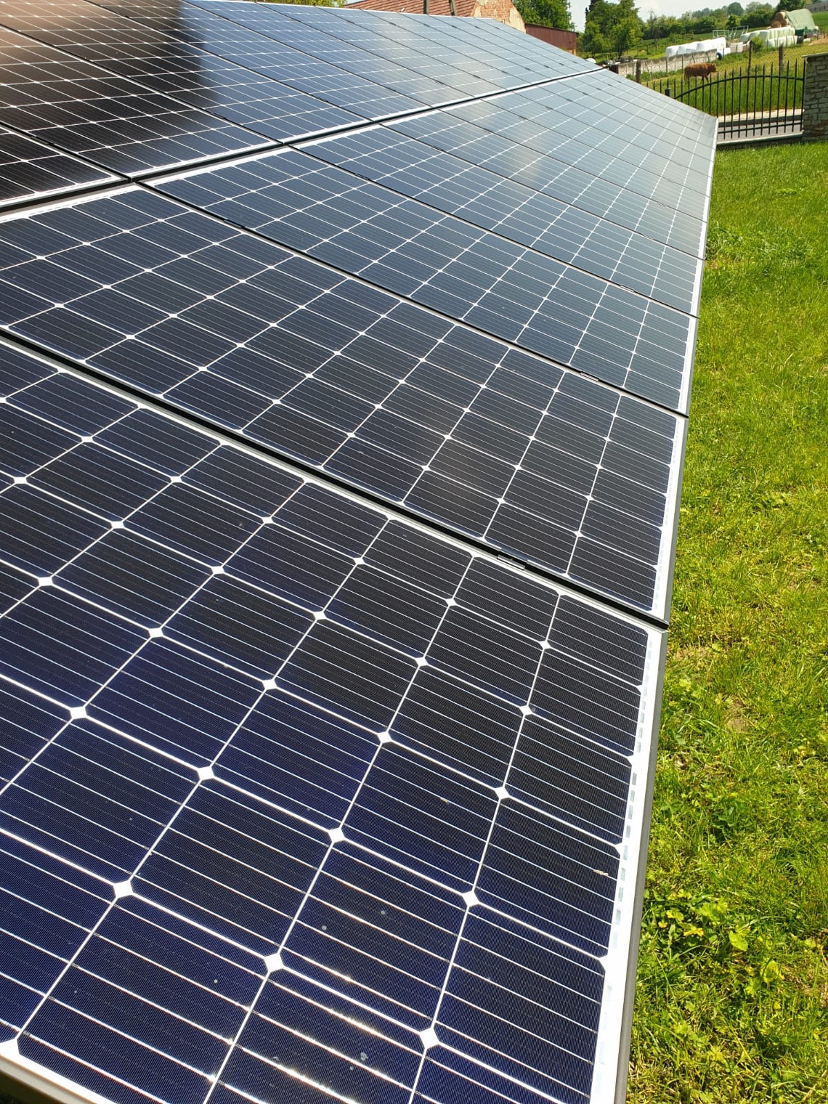 Rząd paneli słonecznych na trawniku, widoczne odbicia w panelach, w tle ogrodzenie posesji.