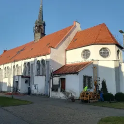 Prace geotechniczne przy zabytkowym kościele z czerwoną dachówką; żółty wiertniczy sprzęt na trawniku, dwóch pracowników w roboczych ubraniach, boczny widok na budynek
