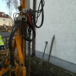 Żółta wiertnica hydrauliczna z wiertłem ślimakowym przy ścianie budynku, obok leży dodatkowe wiertło i łopata.