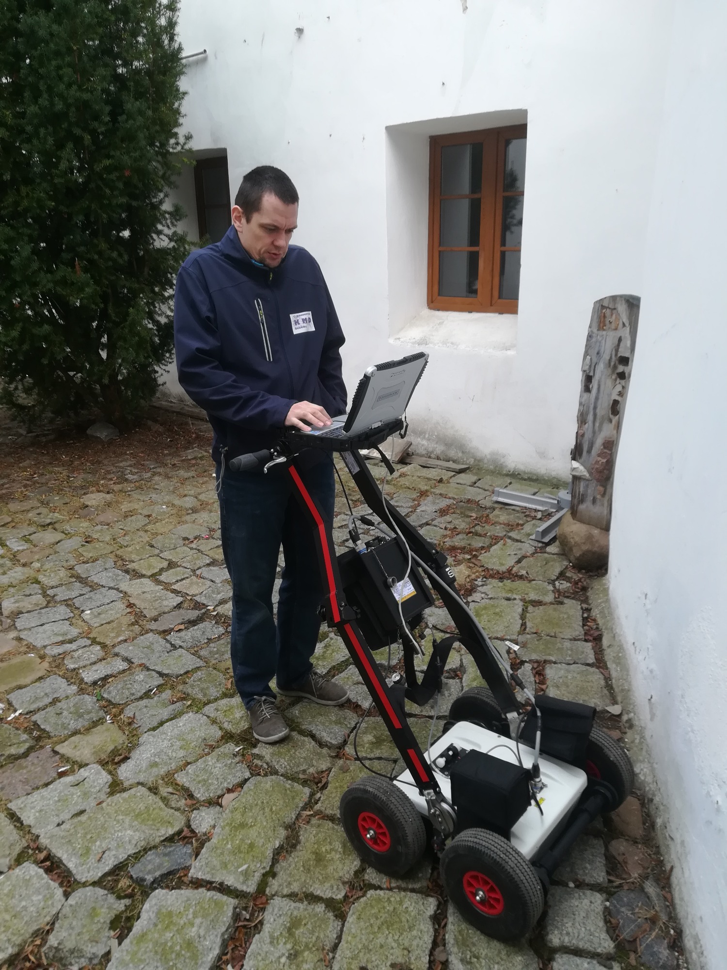 Mężczyzna obsługuje georadar na wózku z laptopem, badanie gruntu na brukowanym dziedzińcu przy białej ścianie budynku z drewnianym oknem.