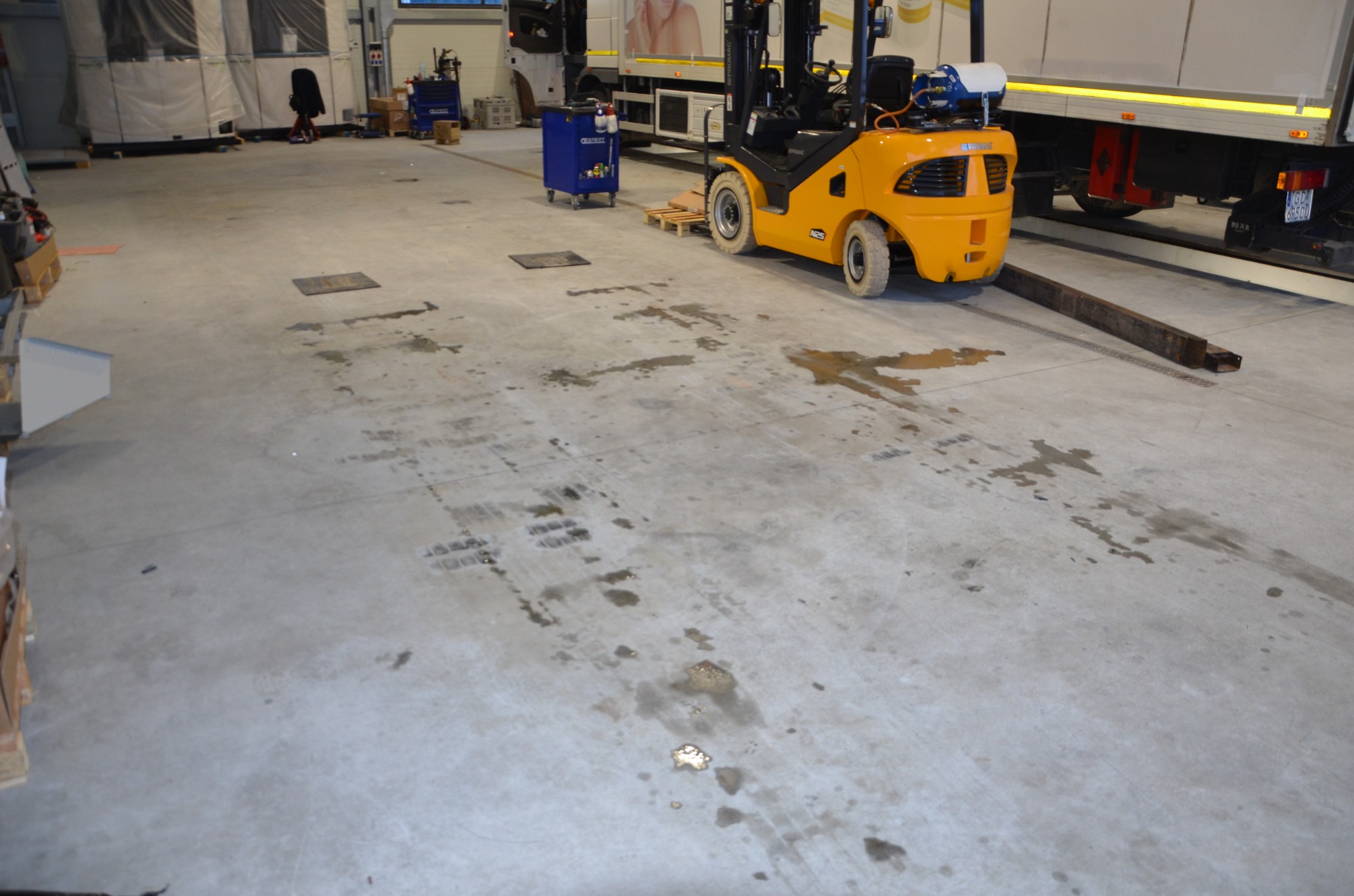 Żółty wózek widłowy na betonowej posadzce z plamami w hali magazynowej, obok zaparkowana ciężarówka.