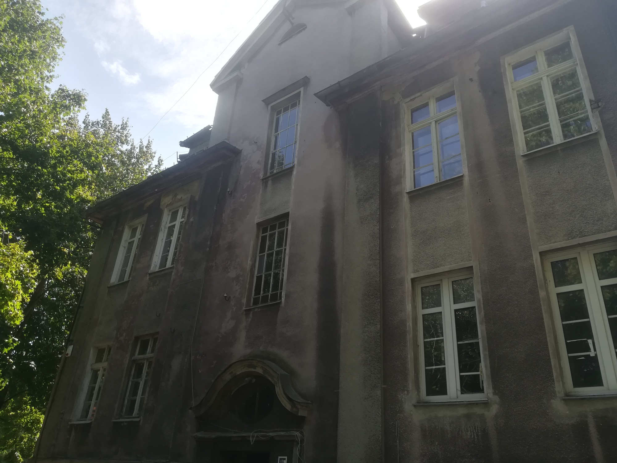 Fasada starego, zaniedbanego budynku z widocznymi śladami wilgoci i braku renowacji, z drewnianymi oknami i dekoracyjnym portalem wejściowym oraz bujną zielenią drzew w tle.