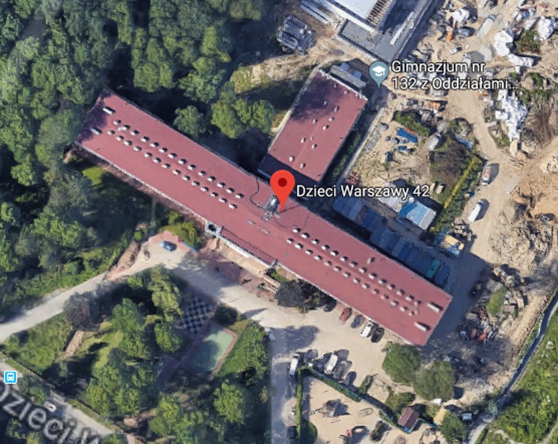 Widok z lotu ptaka na budynek szkoły z czerwonym dachem, otoczony zielenią i parkingiem, z zaznaczoną lokalizacją na mapie.