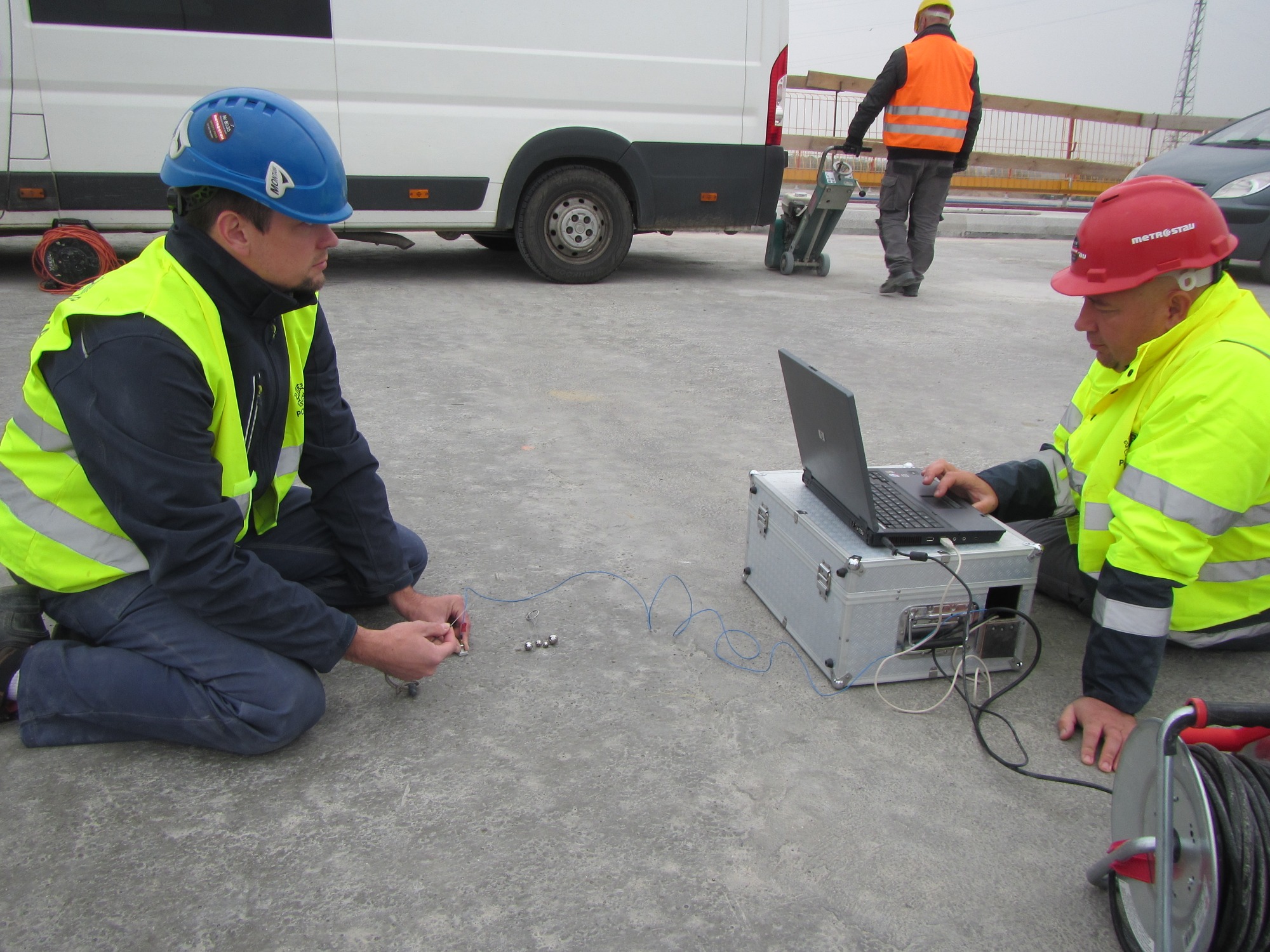 Dwóch inżynierów w Ożarowie Mazowieckim, w kaskach i kamizelkach odblaskowych, analizuje dane pomiarowe na laptopie stojącym na metalowej skrzynce, podczas gdy w tle inny pracownik używa szlifierki...