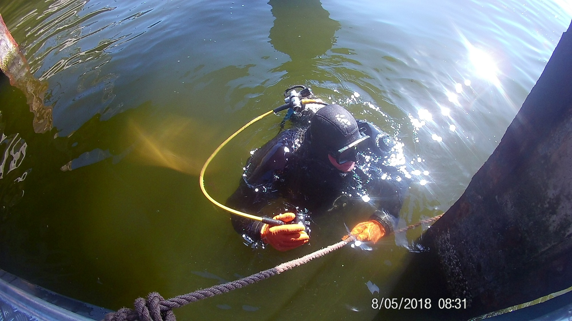 Nurek w ciemnym kombinezonie i pomarańczowych rękawicach trzyma linę w mulistej wodzie, widoczny fragment drewnianej konstrukcji i odbicia słońca na powierzchni.