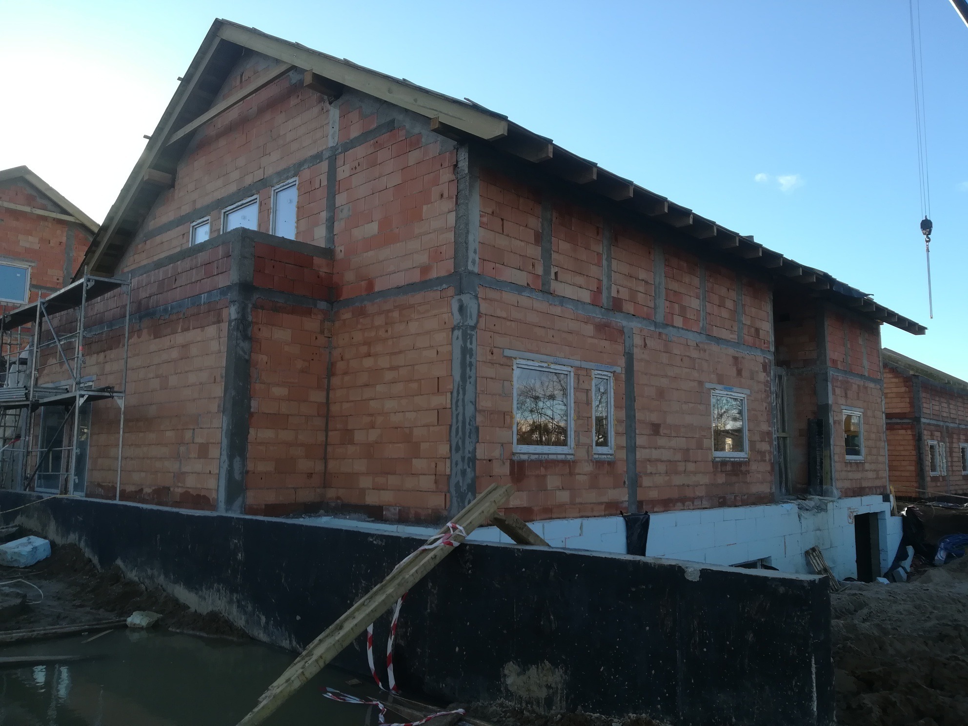 Surowy stan budynku z czerwonej cegły z widocznymi ramami okiennymi, fundamentami i elementami konstrukcyjnymi dachu, na tle błękitnego nieba i częściowo zalanego terenu.