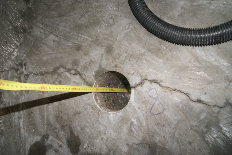 Pomiar średnicy otworu w betonowej posadzce z widocznymi pęknięciami, wykonany żółtą miarką zwijaną, obok widoczny odkurzacz przemysłowy i odręczny napis '8'.