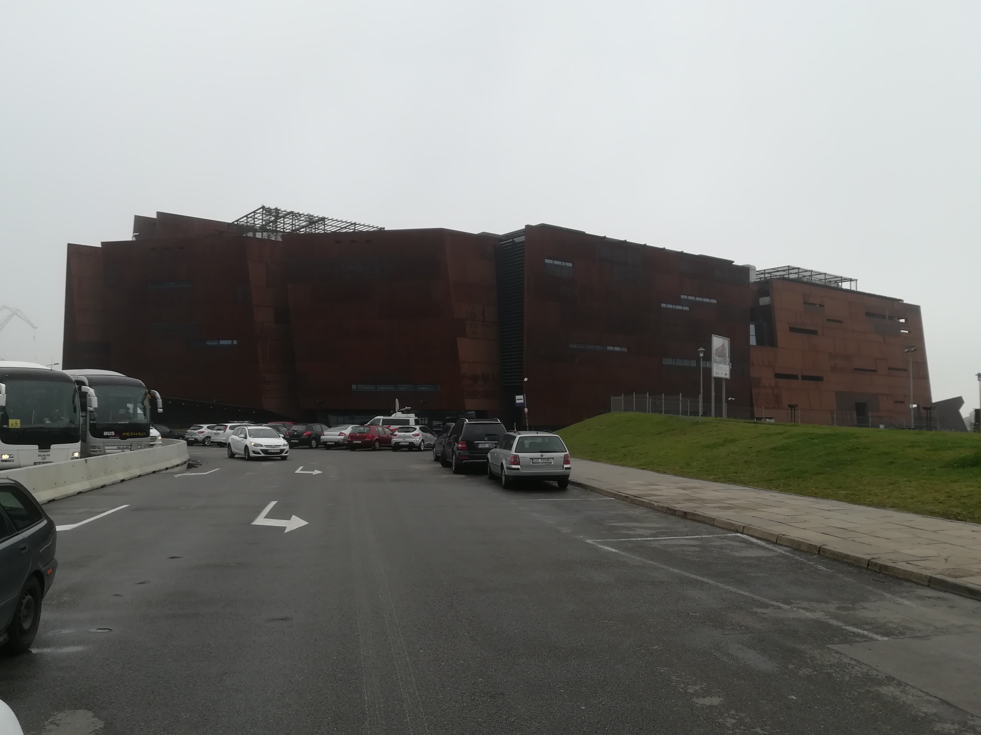 Nowoczesny budynek o rdzawym wykończeniu z metalową konstrukcją na dachu, widoczny z parkingu z samochodami i autobusami w pochmurny dzień.
