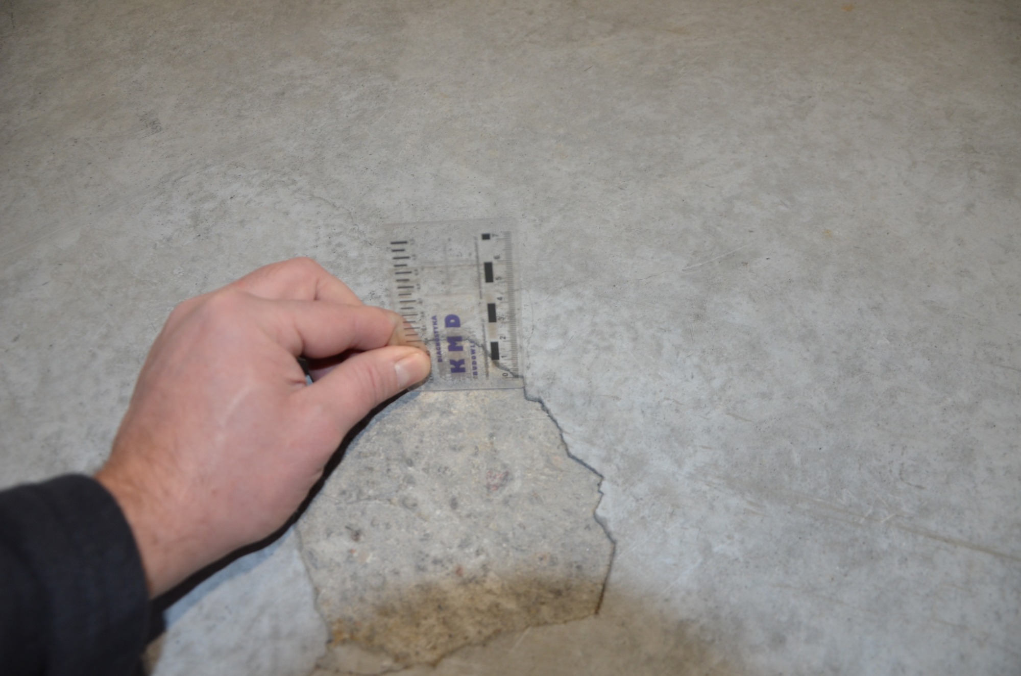 Pomiar linijką uszkodzenia betonowej posadzki, widoczne odpryski i pęknięcia.