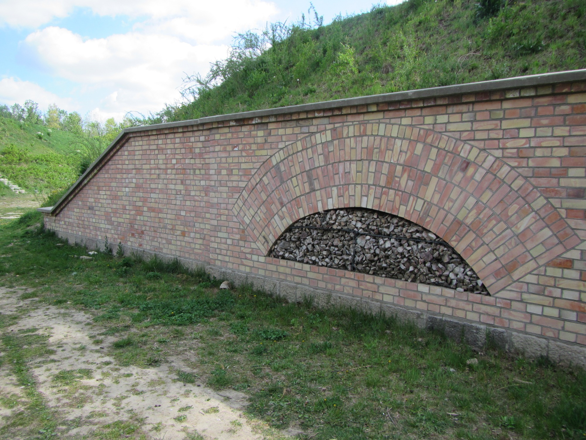 Mur oporowy z cegły klinkierowej z półkolistym otworem wypełnionym kamieniami, zabezpieczający skarpę porośniętą zielenią.