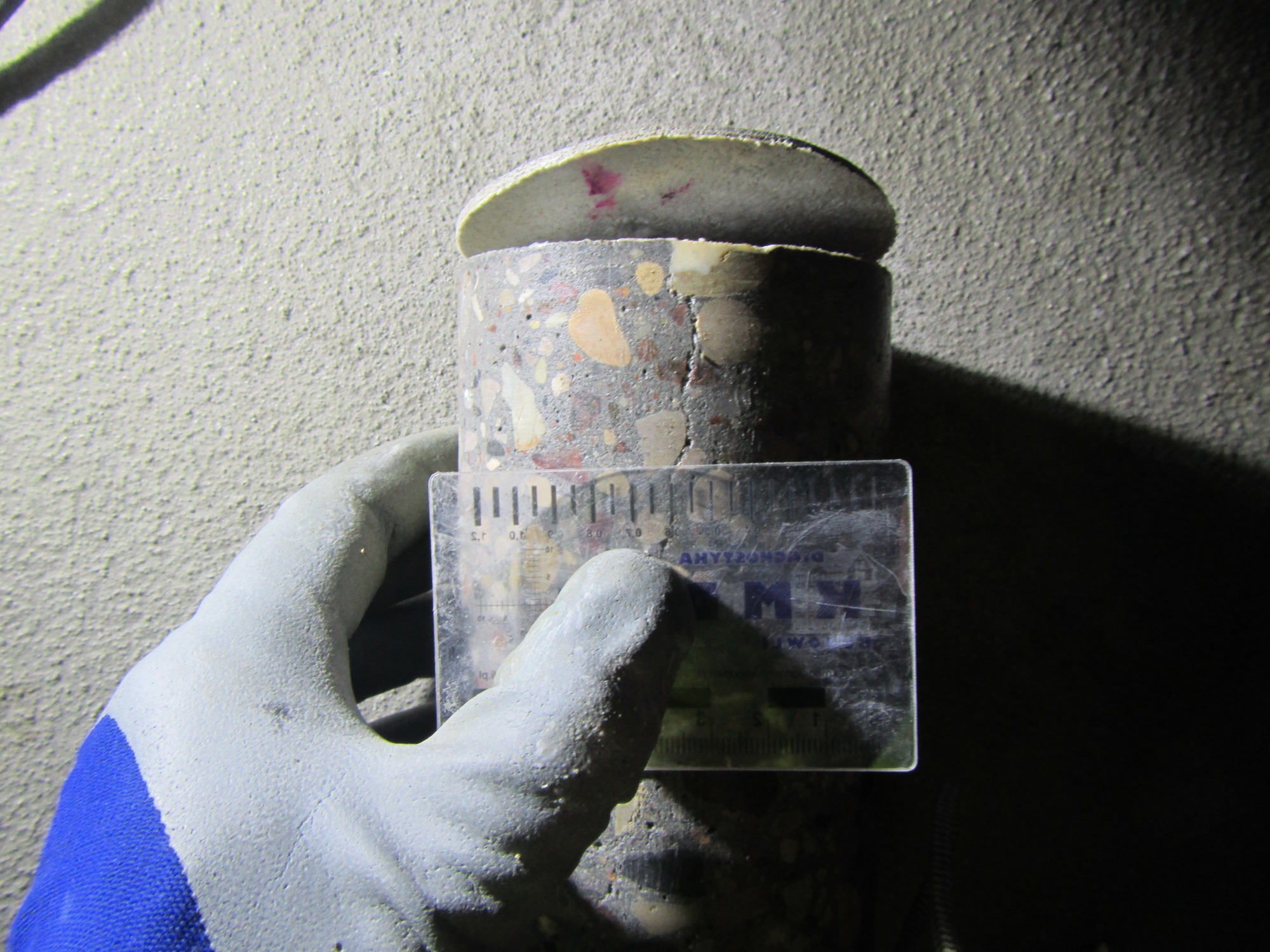 Rdzeń betonowy z widocznymi kruszywami, częściowo odłamany, mierzony przez osobę w rękawicy roboczej za pomocą przezroczystej linijki na tle szarej, teksturowanej ściany.