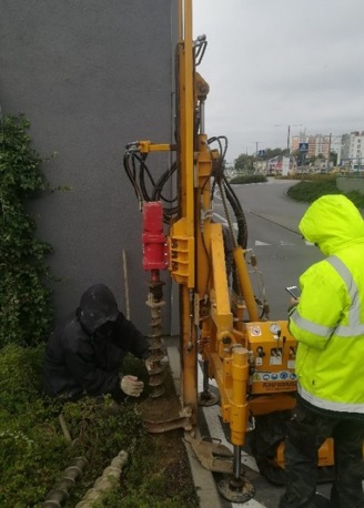 Prace ziemne wykonywane w pobliżu budynku za pomocą wiertnicy z żółtą obudową, obsługiwanej przez dwóch pracowników w odzieży ochronnej.