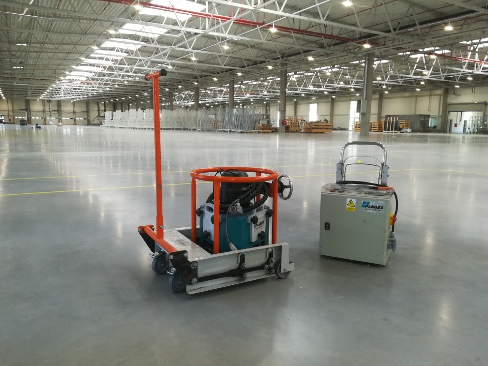 Maszyny do obróbki posadzek przemysłowych w przestronnej hali magazynowej z widoczną konstrukcją dachu.