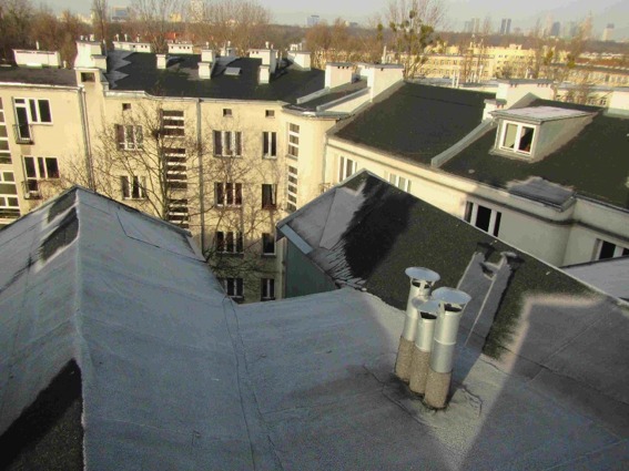 Widok z góry na płaski dach budynku z trzema kominami wentylacyjnymi, w tle budynki mieszkalne i drzewa bez liści.