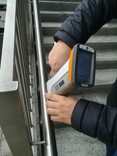 Osoba w kurtce dokonuje pomiaru metalowej barierki schodowej za pomocą przenośnego analizatora, wyświetlającego wynik 1.4318.