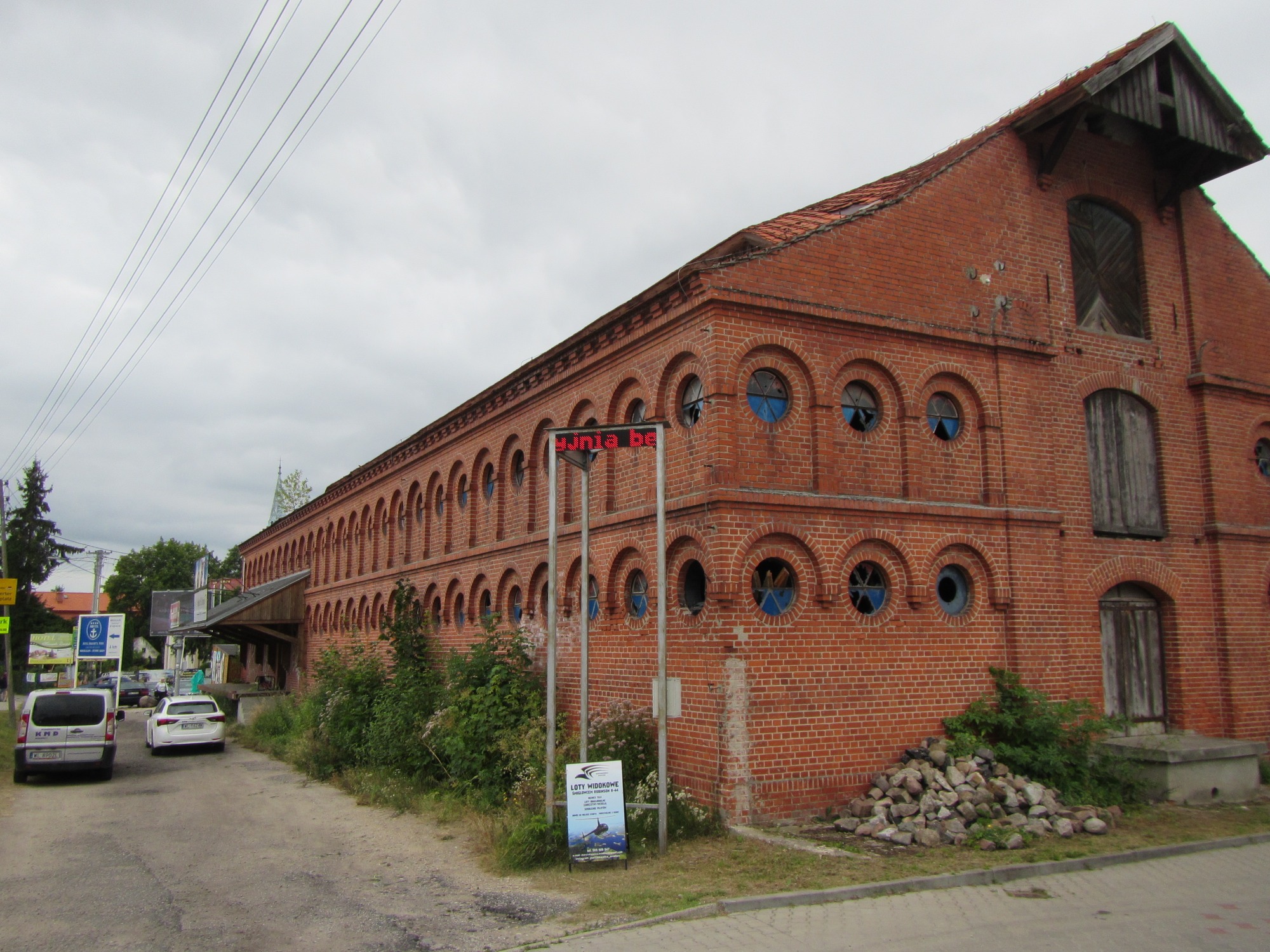 Ceglany budynek z rzędem okrągłych okien z niebieskim szkłem, częściowo zniszczonym, z drewnianymi okiennicami na górze, obok droga i zaparkowane samochody, widoczny szyld reklamowy.